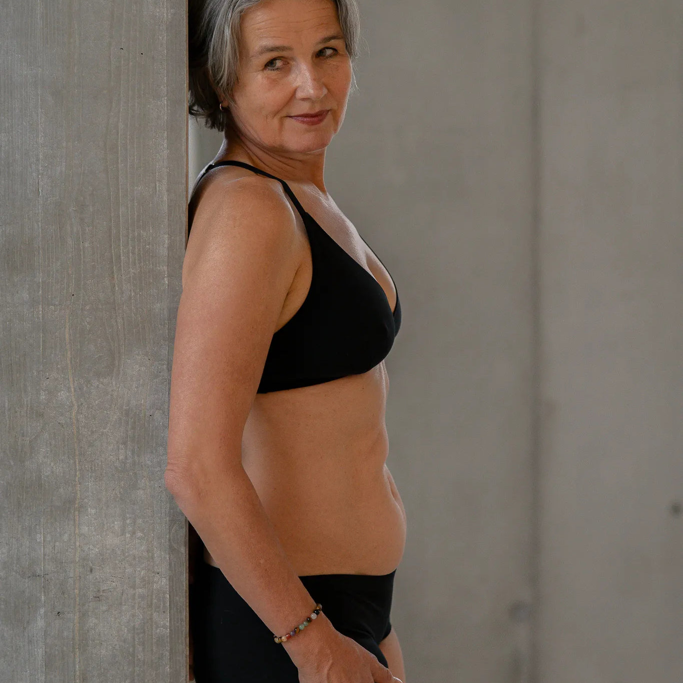 Model steht gegen eine Betonsäule gelehnt, die Hände lässig auf dem Oberschenkel. Sie trägt das Dina Bralette und die Wilma Panty und blickt an der Kamera vorbei.