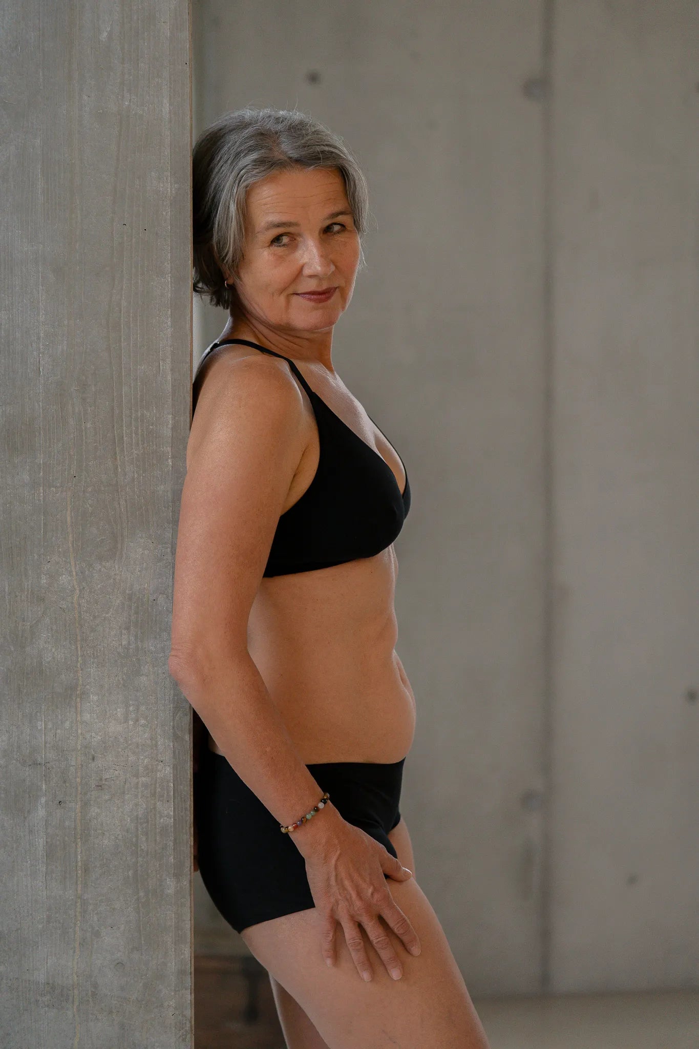 Model steht gegen eine Betonsäule gelehnt, die Hände lässig auf dem Oberschenkel. Sie trägt das Dina Bralette und die Wilma Panty und blickt an der Kamera vorbei.