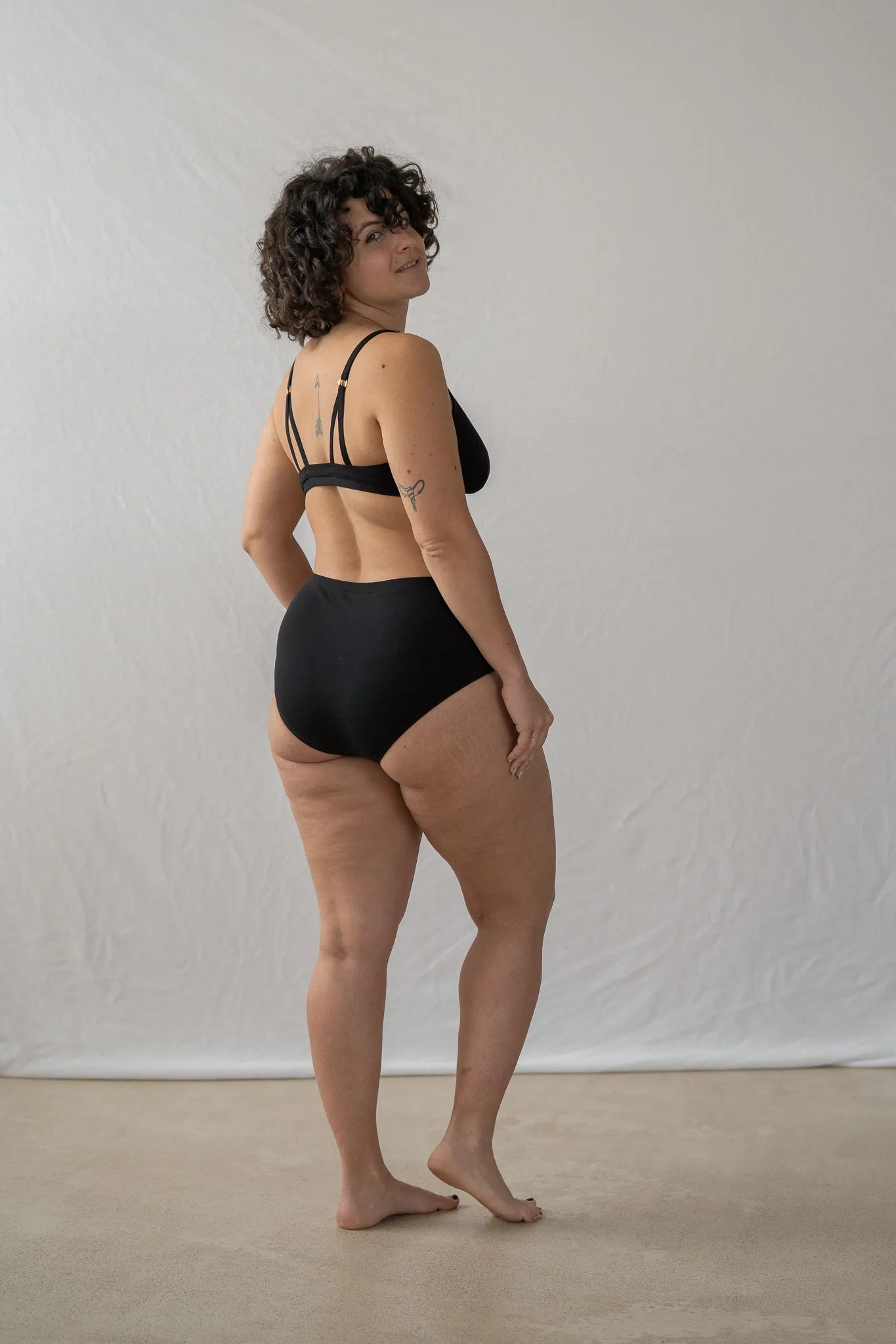 Model steht mit dem Rücken zur Kamera vor einem weissen Hintergrund. Sie blickt über die Schulter in die Kamera und trägt das Dina Bralette und den Odette High Waist Slip.