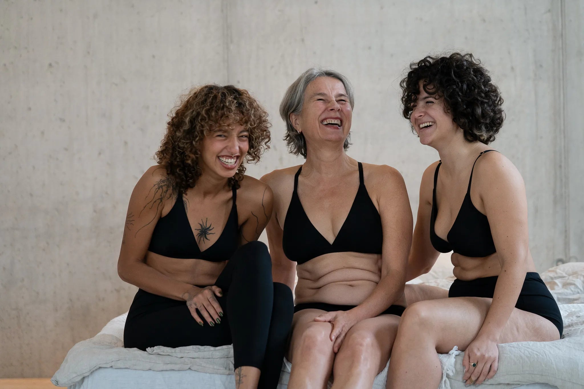 Drei Models sitzen nebeneinander auf der Bettkante und lachen zusammen. Sie tragen unsere neue Cotton Collection, bestehend aus dem Dina Bralette, der Inge Leggins, der Wilma Panty und dem Odette High Waist Slip.