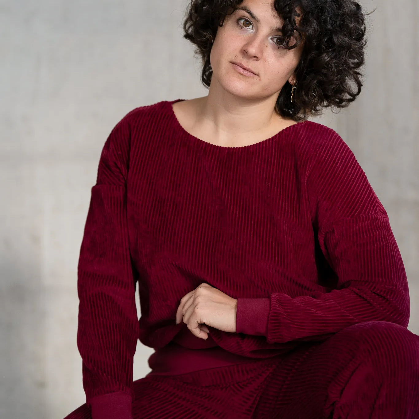 Model kniet am Boden, sie trägt den Chloé Pullover und die Fiona Hose in Ruby Red und blickt direkt in die Kamera. Ein Bein hat sie aufgestellt.