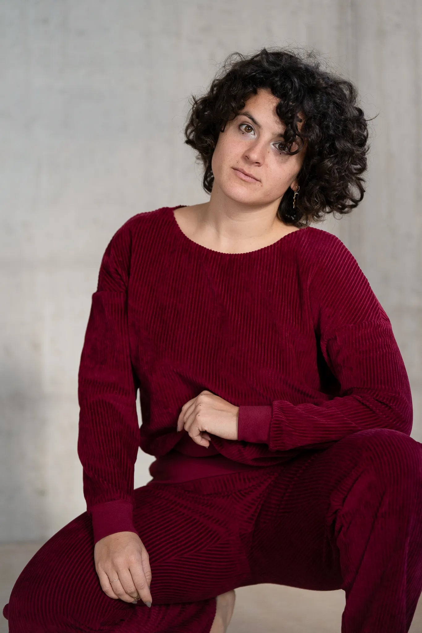 Model kniet am Boden, sie trägt den Chloé Pullover und die Fiona Hose in Ruby Red und blickt direkt in die Kamera. Ein Bein hat sie aufgestellt.