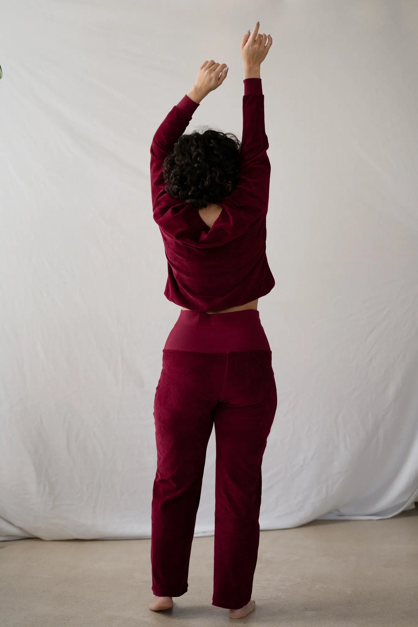 Model steht mit dem Rücken zur Kamera, die Hände hat sie in die Luft gestreckt. Sie trägt den Chloé Pullover und die Fiona Hose in Ruby Red.
