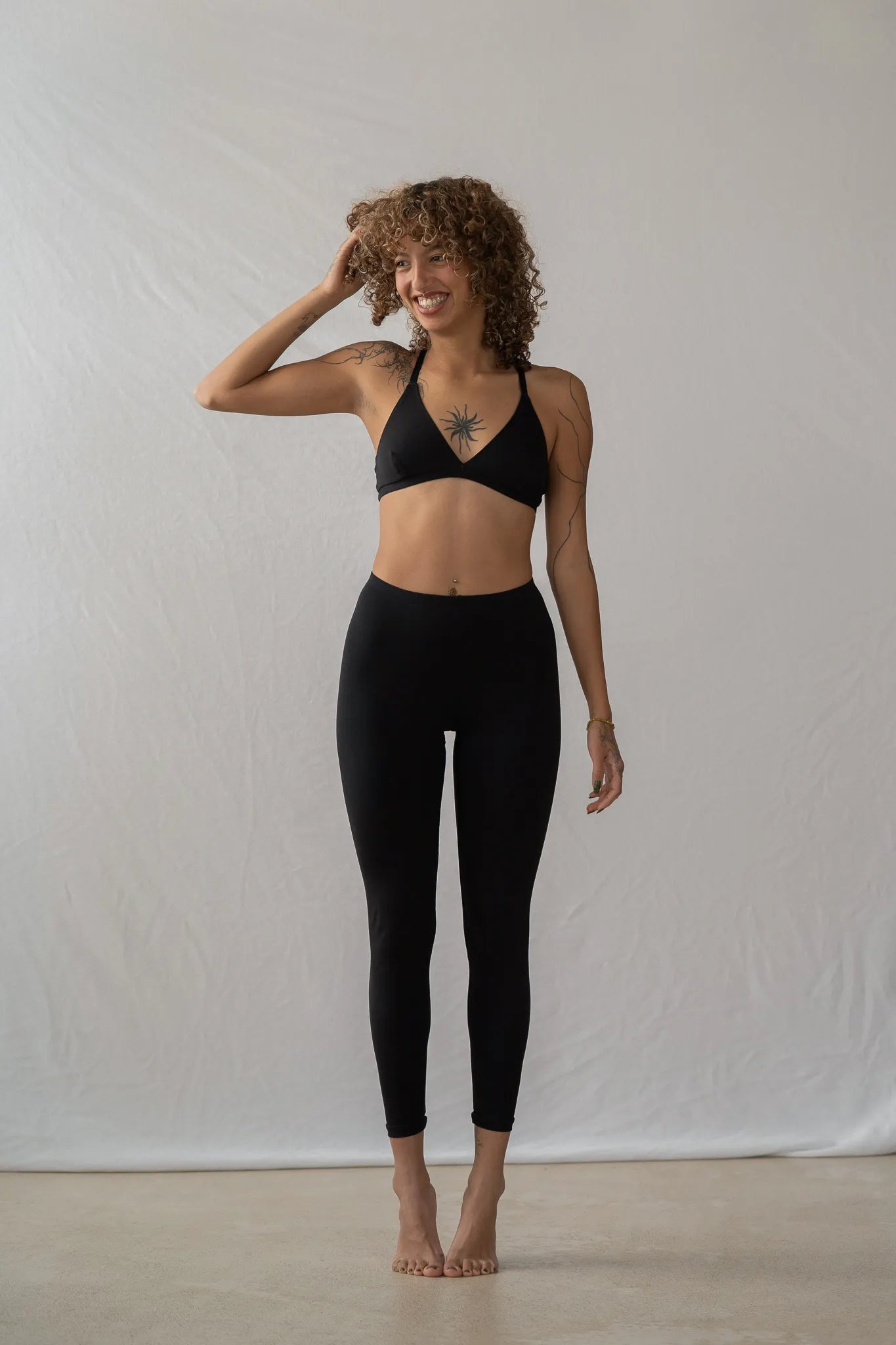 Model steht vor einem weissen Hintergrund auf den Zehenspitzen und lächelt. Sie trägt die Inge Leggins und das Dina Bralette.