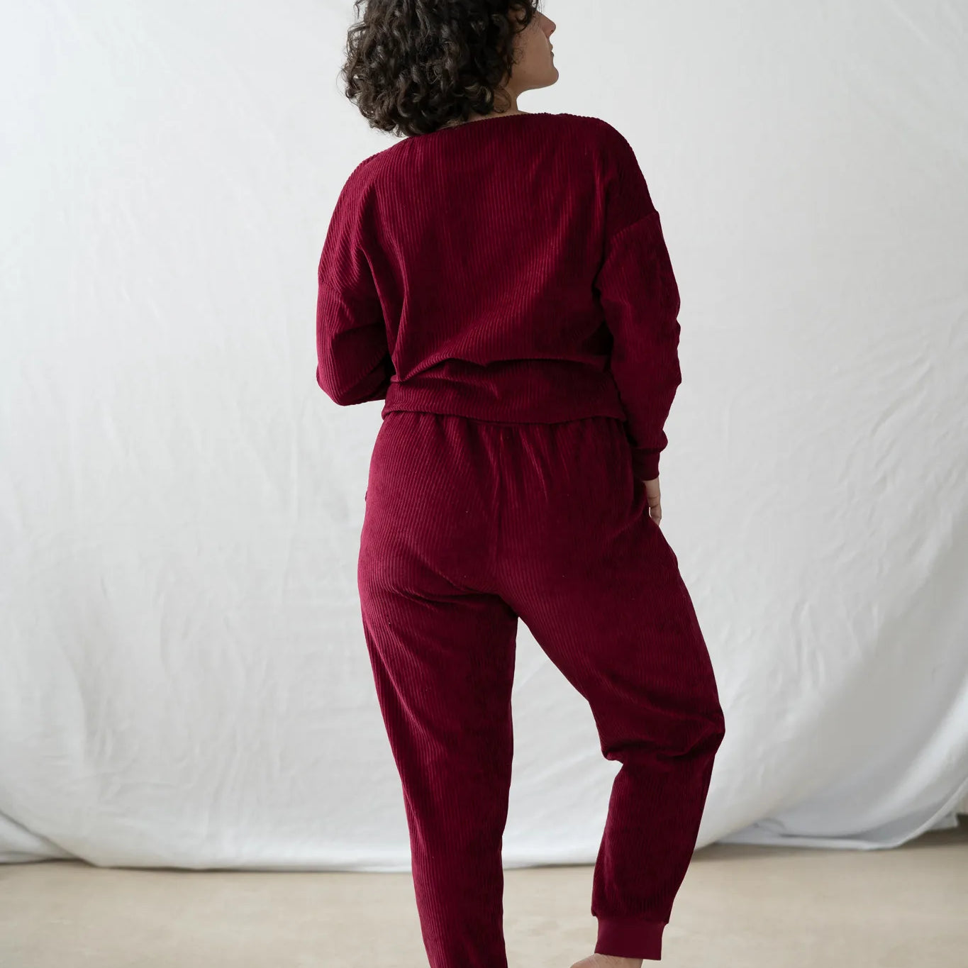 Model steht mit dem Rücken zur Kamera gerichtet, den Kopf leicht zur Seite gelegt. Sie trägt den Chloé Pulli und die Kelly Hose in Ruby Red.