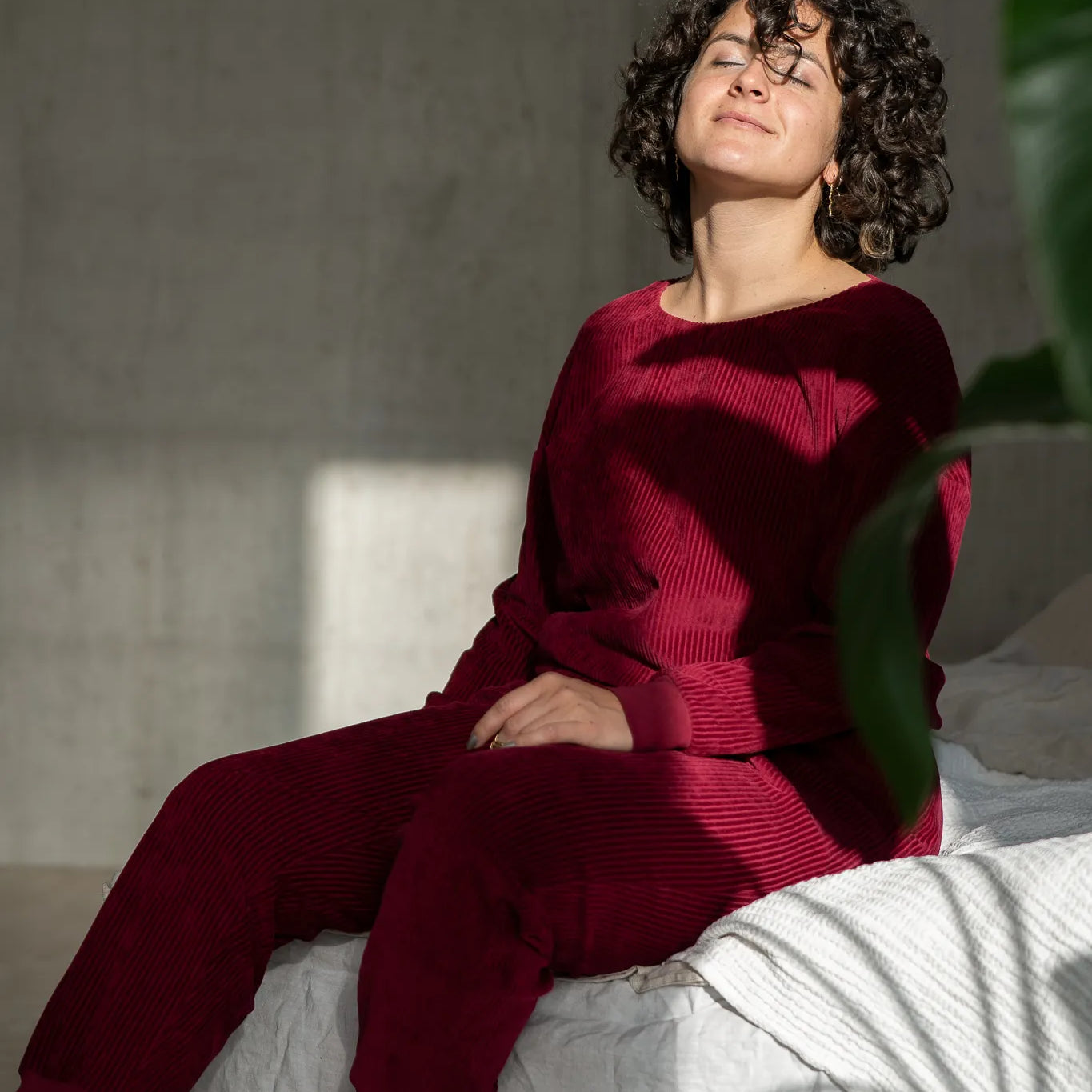 Model sitzt auf der Bettkante und trägt unsere Kelly Pant und den Chloé Pulli in Ruby Red. Die Sonne scheint durchs Fenster in ihr Gesicht.