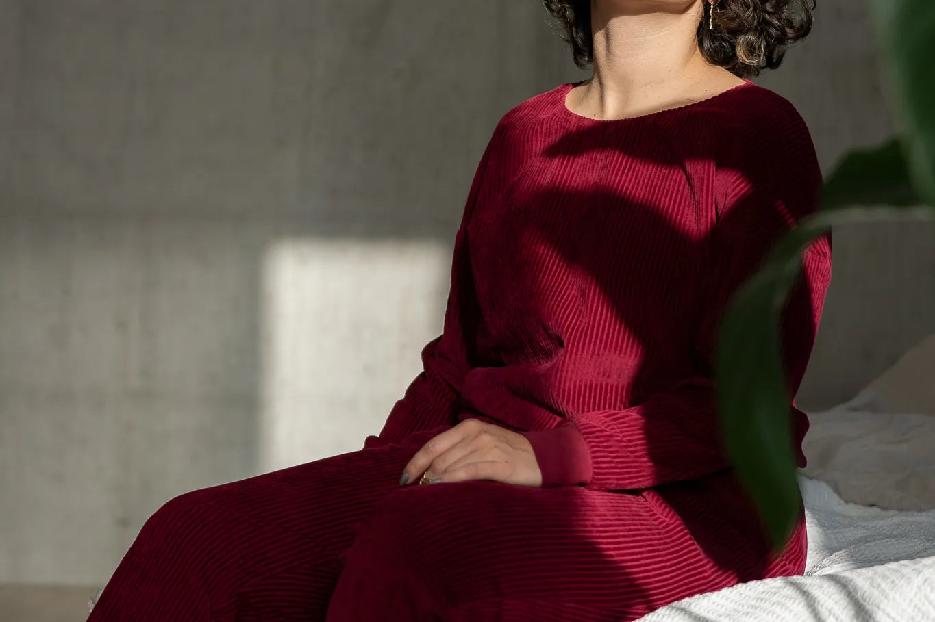 Model sitzt auf der Bettkante und trägt unsere Kelly Pant und den Chloé Pulli in Ruby Red. Die Sonne scheint durchs Fenster in ihr Gesicht.