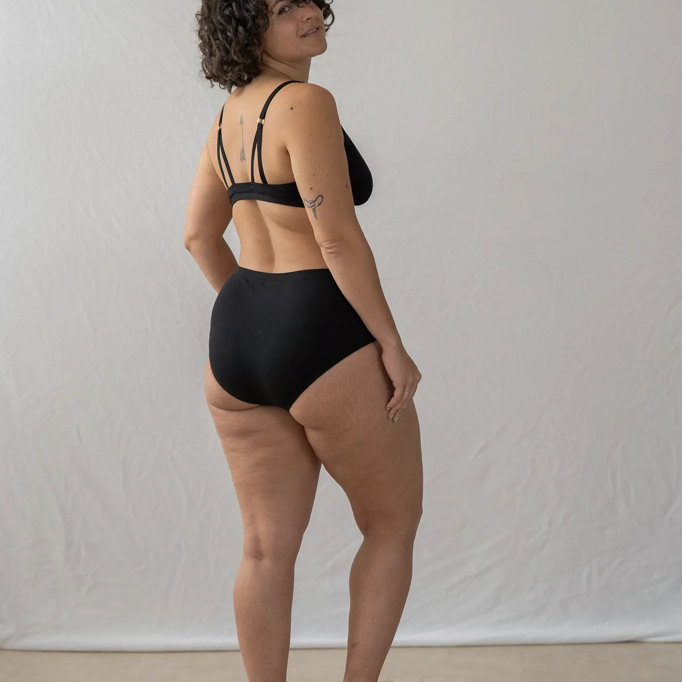 Model steht mit dem Rücken zur Kamera gewandt und wirft einen Blick über die Schulter. Sie trägt das Dina Bralette und den Odette High Waist Slip.