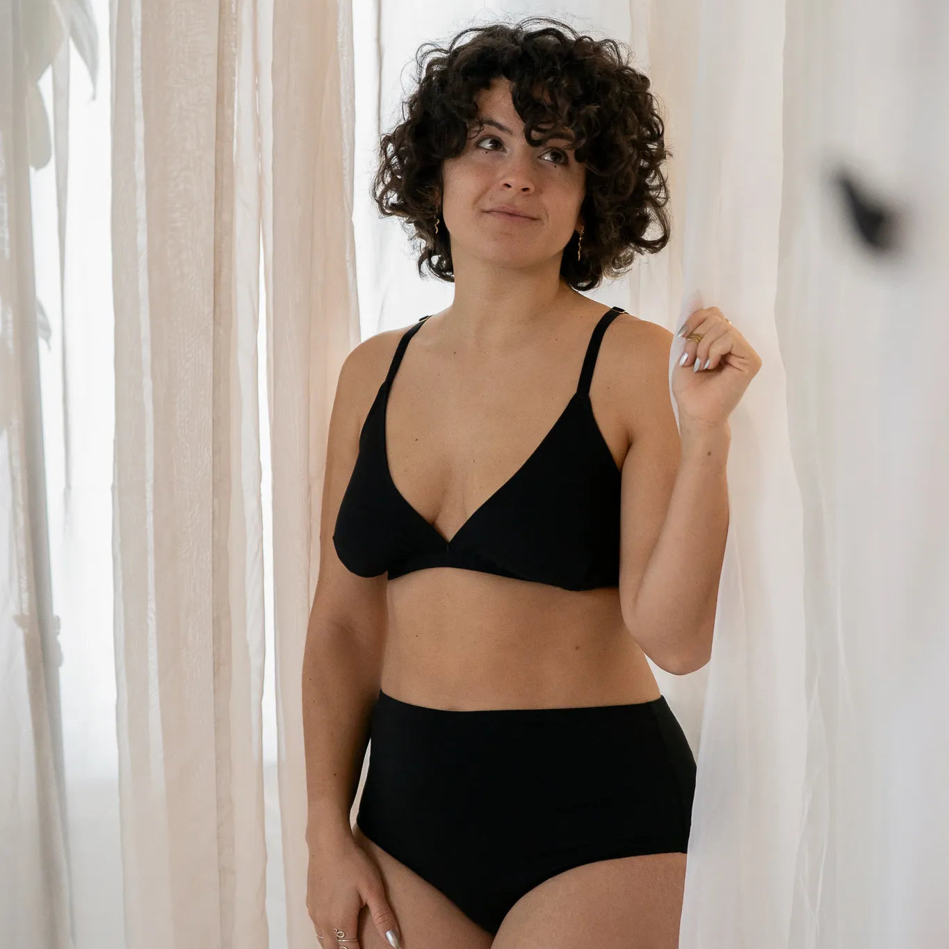 Model steht vor einem grossen Vorhang, das Licht schimmert weich hindurch. Sie trägt das Dina Bralette und den Odette High Waist Slip.