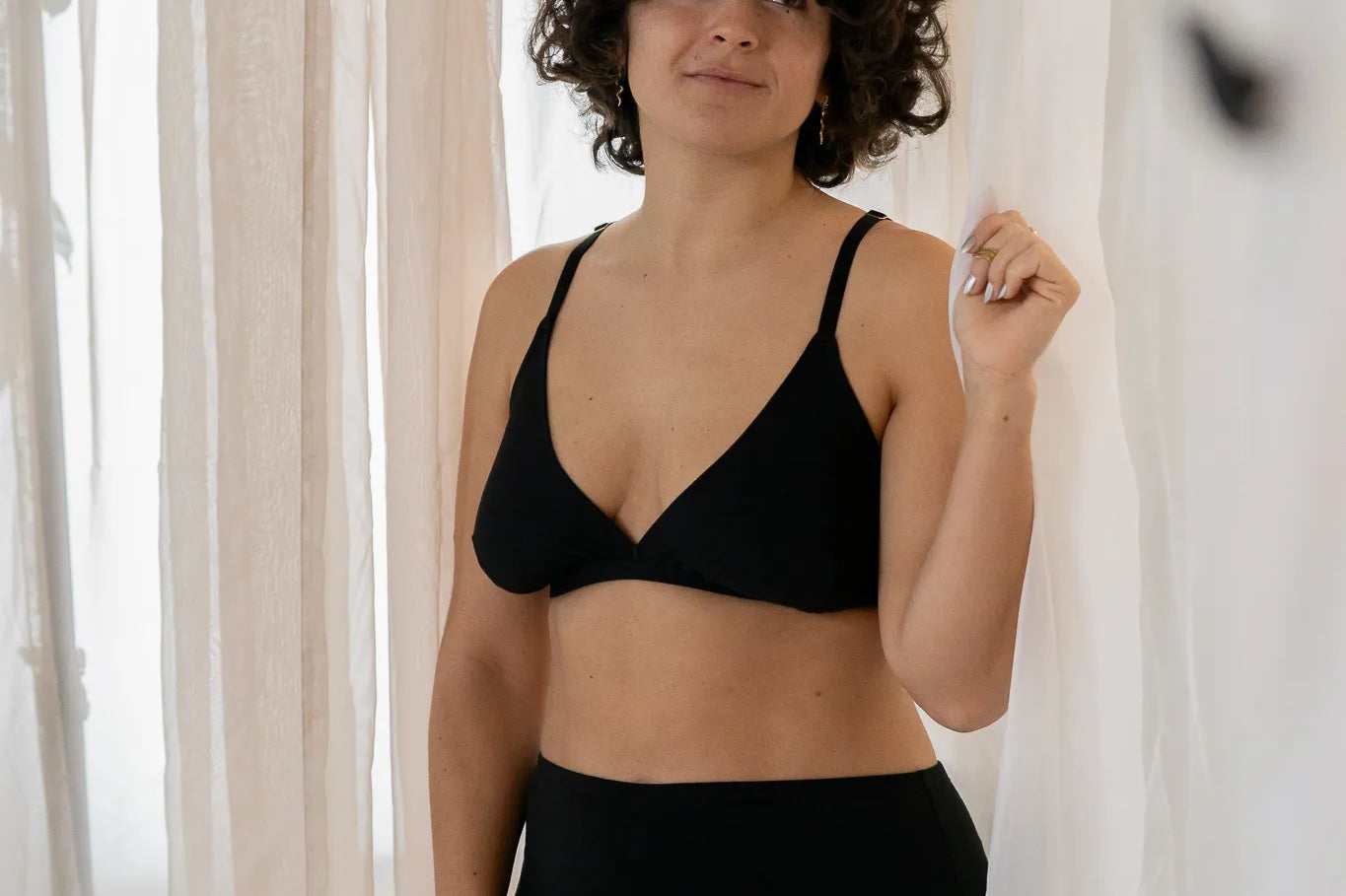 Model steht vor einem grossen Vorhang, das Licht schimmert weich hindurch. Sie trägt das Dina Bralette und den Odette High Waist Slip.