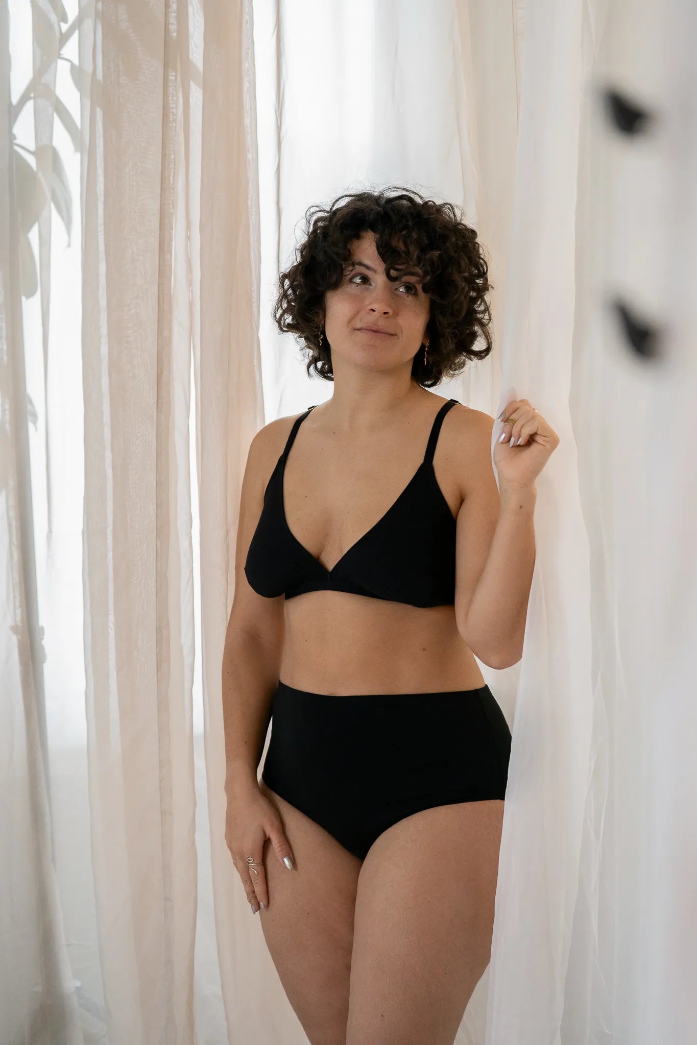 Model steht vor einem grossen Vorhang, das Licht schimmert weich hindurch. Sie trägt das Dina Bralette und den Odette High Waist Slip.