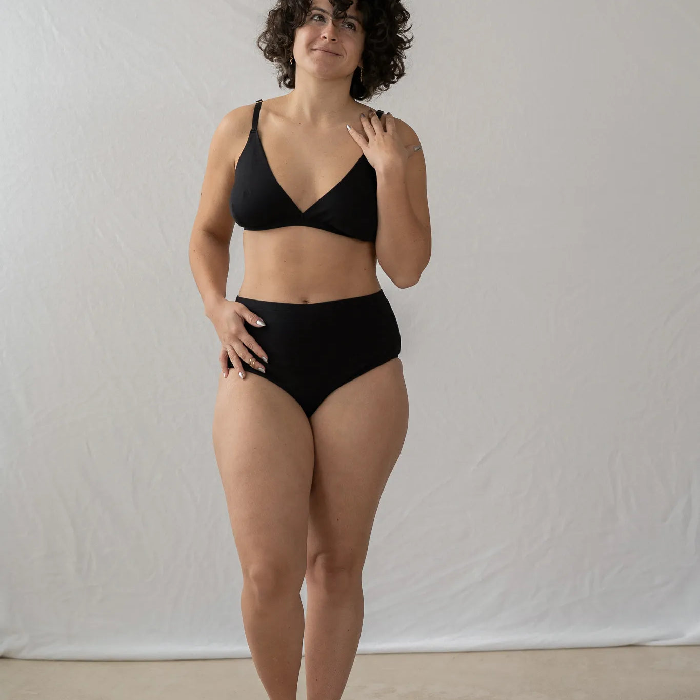 Model steht vor einem weissen Vorhang. Sie trägt das Dina Bralette und den Odette High Waist Slip und lächelt verträumt zur Seite.