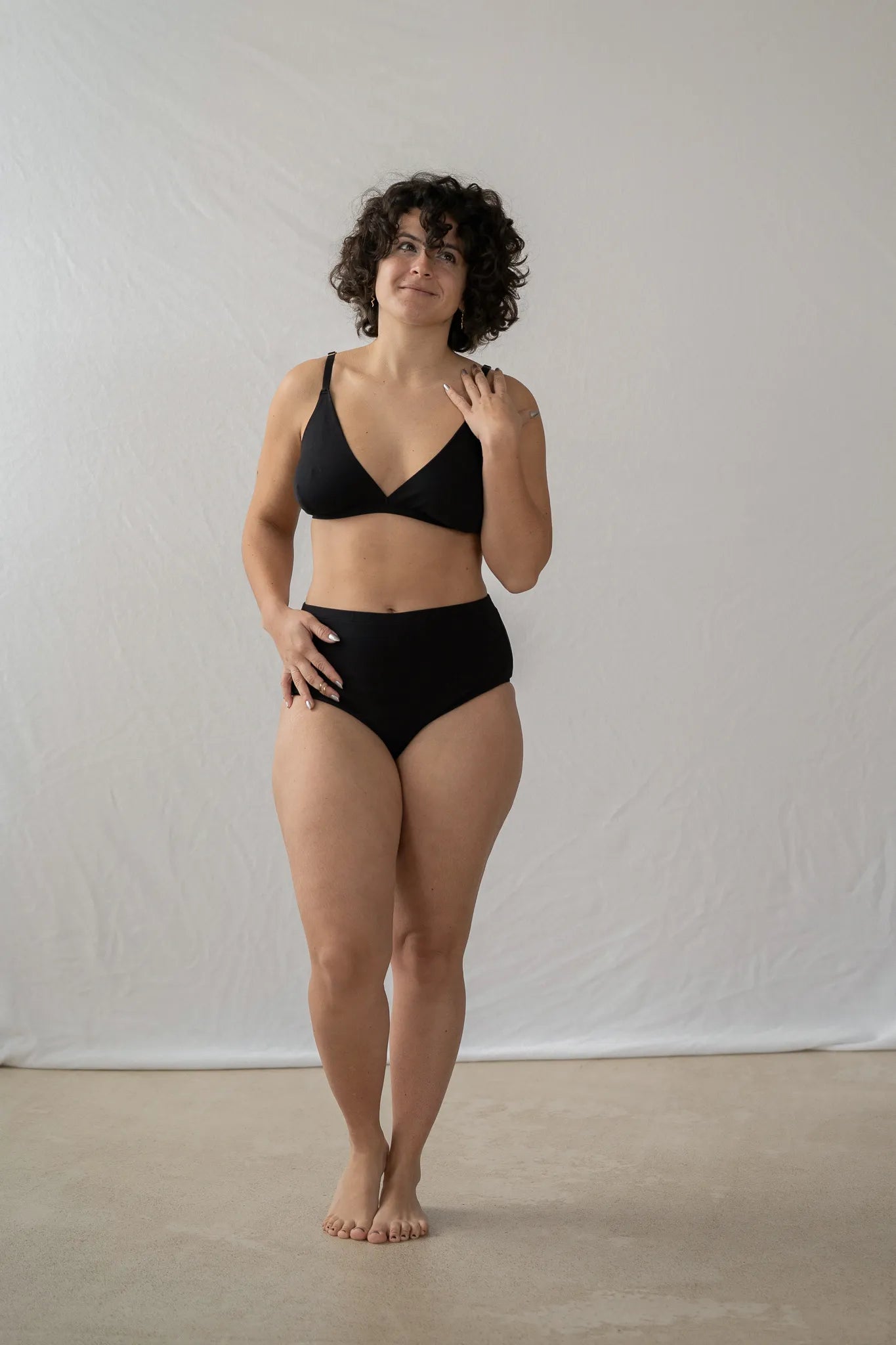 Model steht vor einem weissen Vorhang. Sie trägt das Dina Bralette und den Odette High Waist Slip und lächelt verträumt zur Seite.