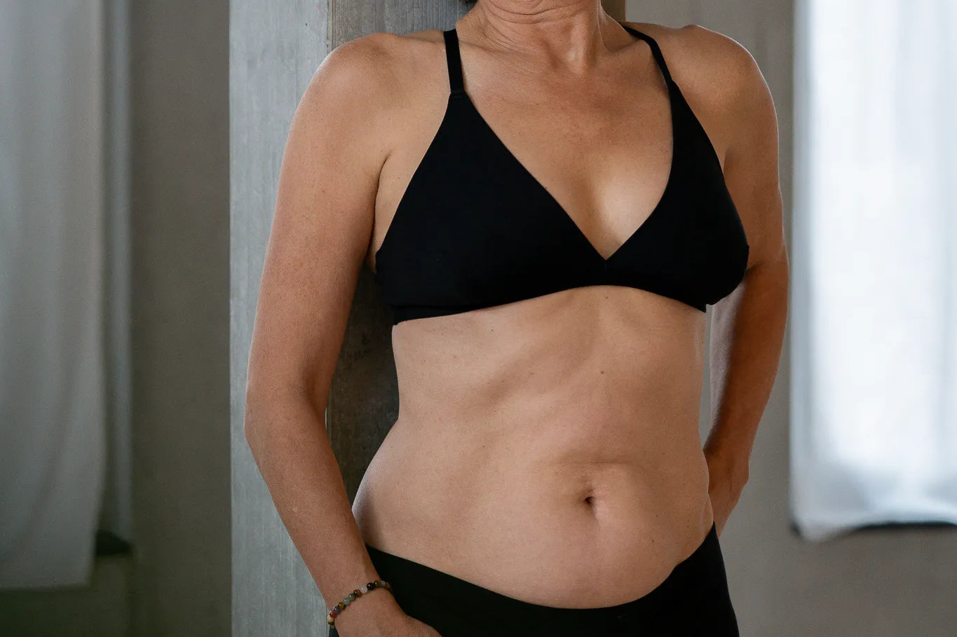 Model steht lässig gegen eine Betonsäule gelehnt. Sie trägt das Dina Bralette und die Wilma Panty und blickt zur Seite.