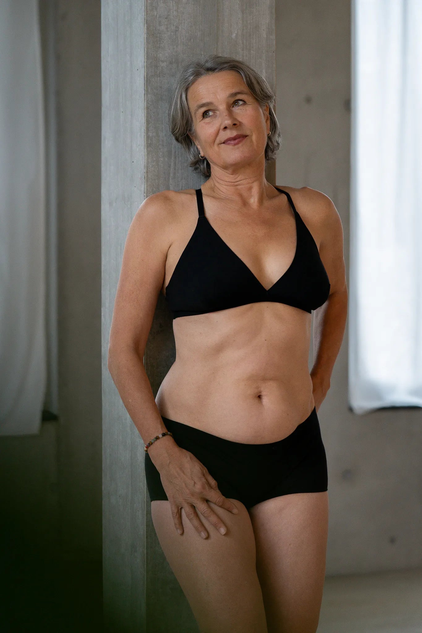 Model steht lässig gegen eine Betonsäule gelehnt. Sie trägt das Dina Bralette und die Wilma Panty und blickt zur Seite.