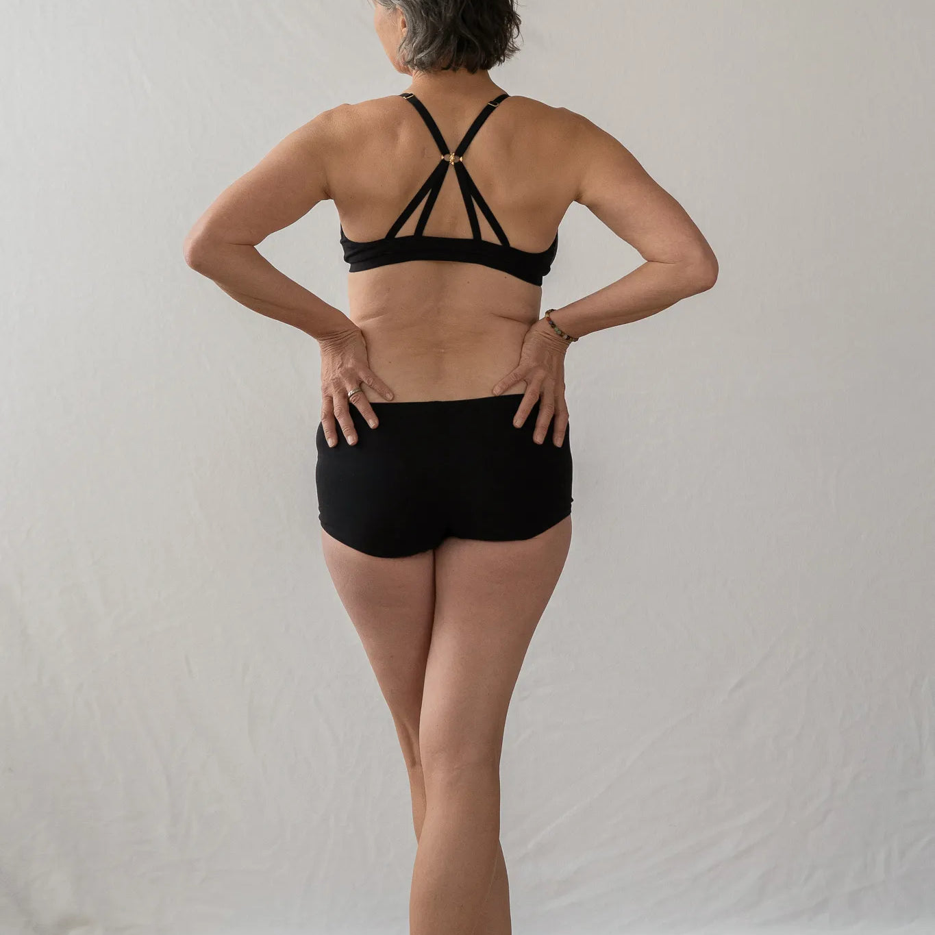 Model steht mit dem Rücken zur Kamera. Sie hat die Hände in die Hüfte gestemmt und blickt leicht seitwärts. Sie trägt das Dina Bralette und die Wilma Panty.