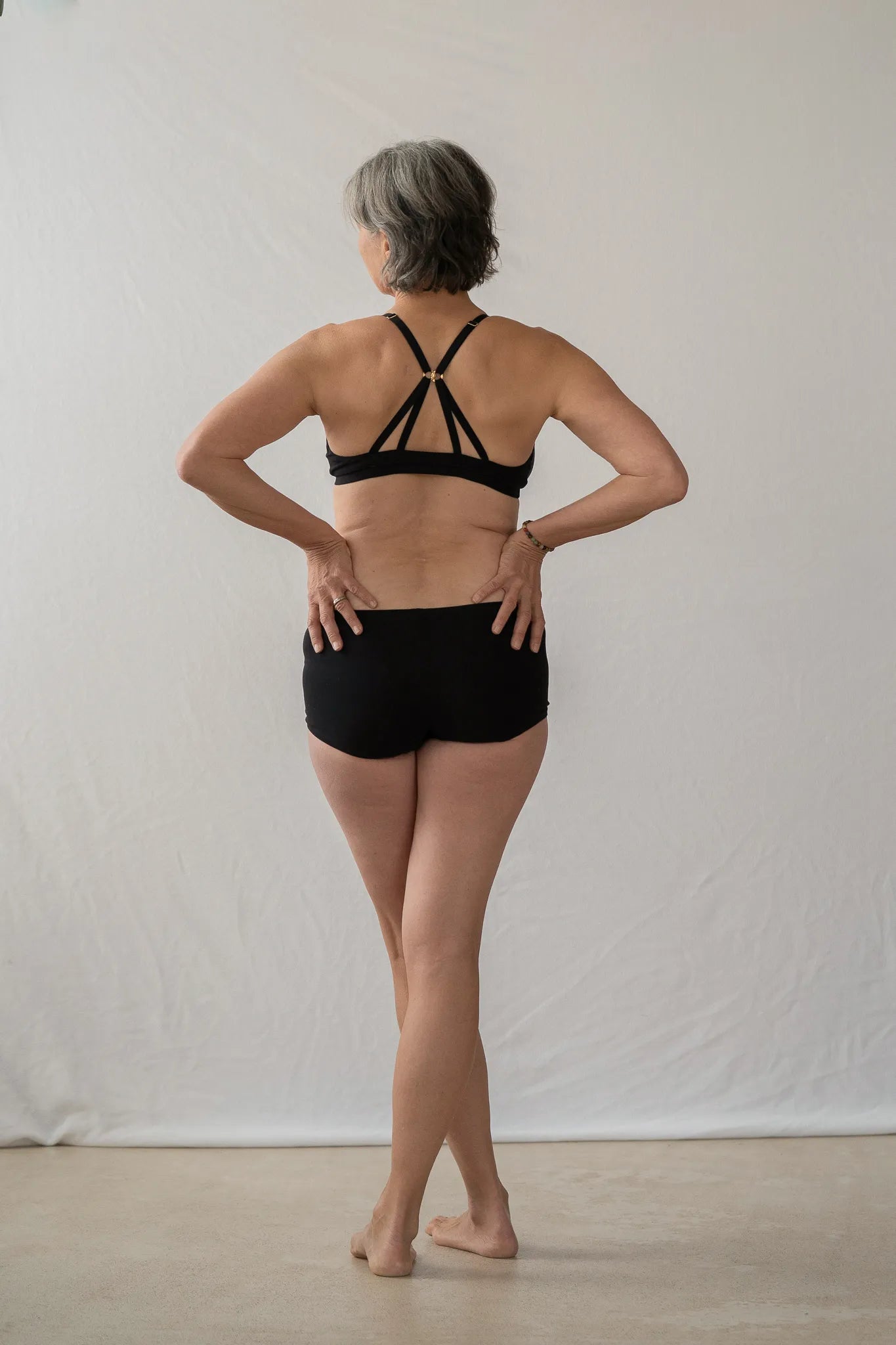 Model steht mit dem Rücken zur Kamera. Sie hat die Hände in die Hüfte gestemmt und blickt leicht seitwärts. Sie trägt das Dina Bralette und die Wilma Panty.