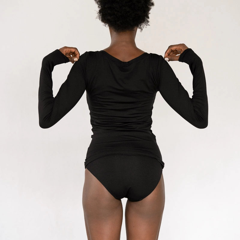 Model steht vor weissem Hintergrund, den Rücken zur Kamera. Sie trägt das Dolly Longsleeve in Schwarz und den Thea Slip.