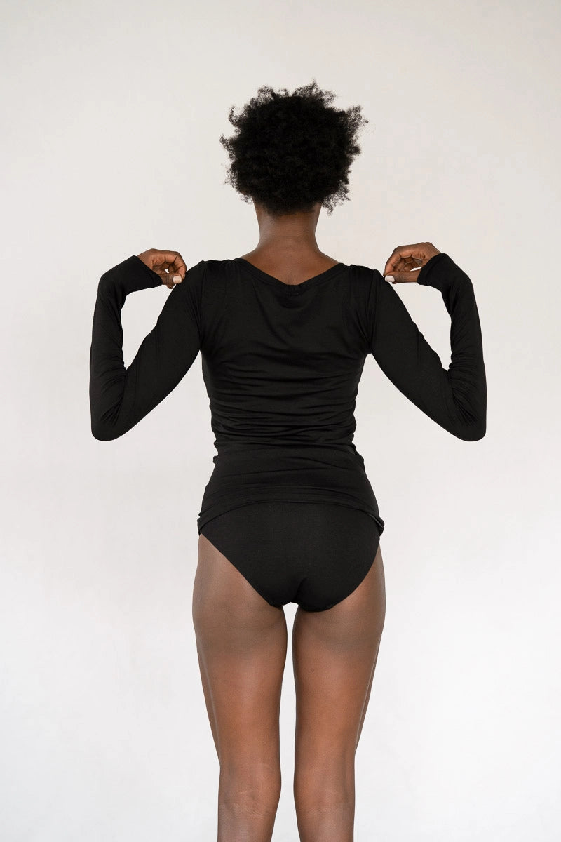 Model steht vor weissem Hintergrund, den Rücken zur Kamera. Sie trägt das Dolly Longsleeve in Schwarz und den Thea Slip.