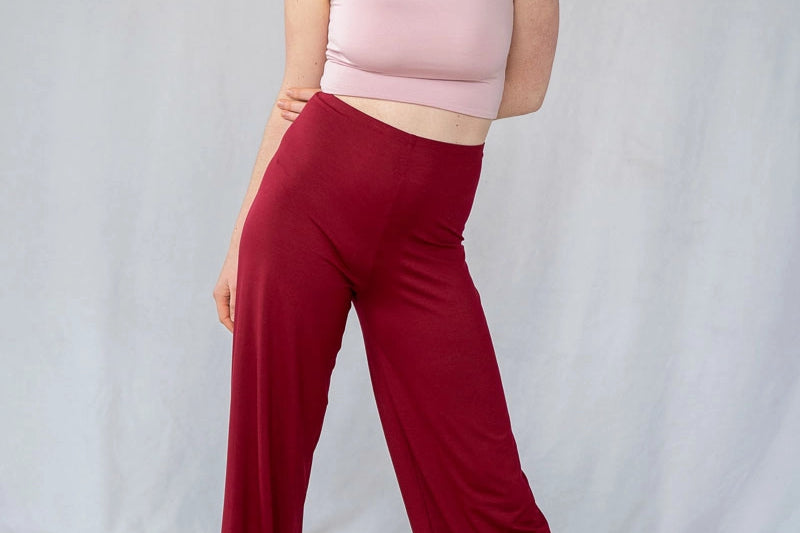 Model steht vor weissem Hintergrund, breitbeinig und das Gewicht auf ein Bein verlagert. Sie trägt das Carla Top in Dahlia Rose und die Renée Hose in der Farbe Ruby Red.