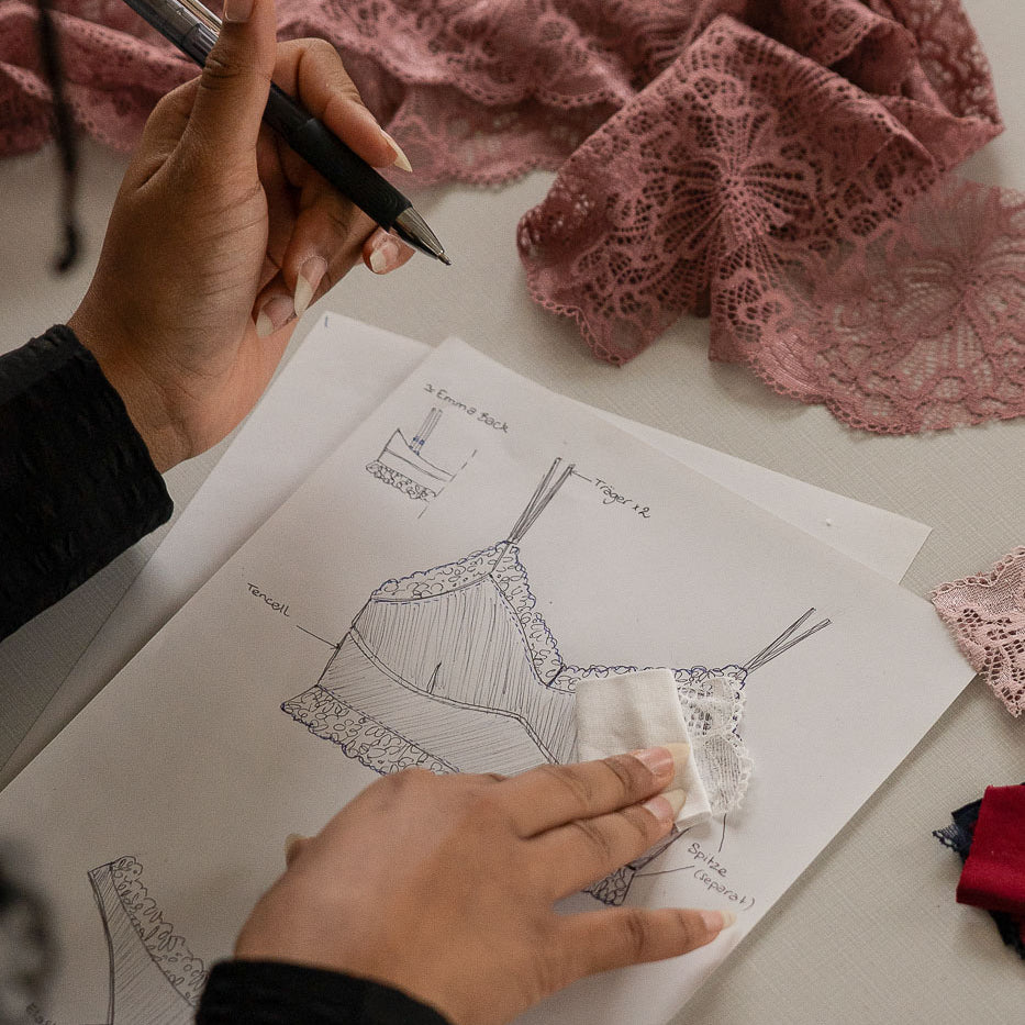 Mathilda sitzt an einem Lingeriedesign, das sie mit Stift auf Papier gezeichnet hat. Rund um das Papier liegt Spitze in verschiedenen Farben.