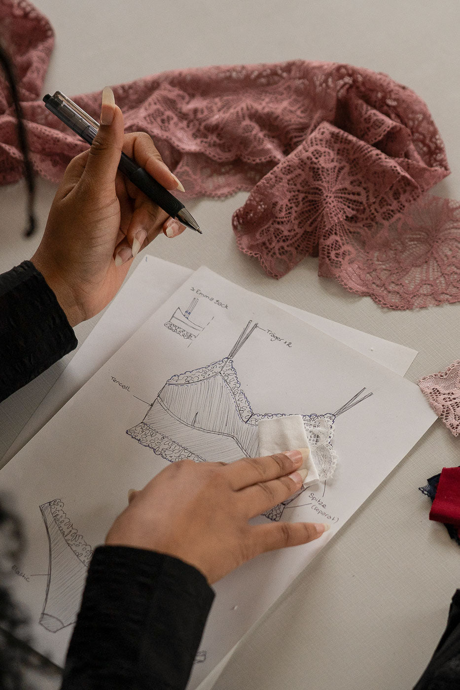 Mathilda sitzt an einem Lingeriedesign, das sie mit Stift auf Papier gezeichnet hat. Rund um das Papier liegt Spitze in verschiedenen Farben.