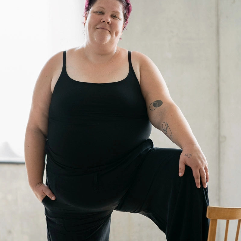 Model steht mit einem Bein auf einem Holzstuhl. Sie trägt das Carla Camitop und die Renée Hose in Schwarz.