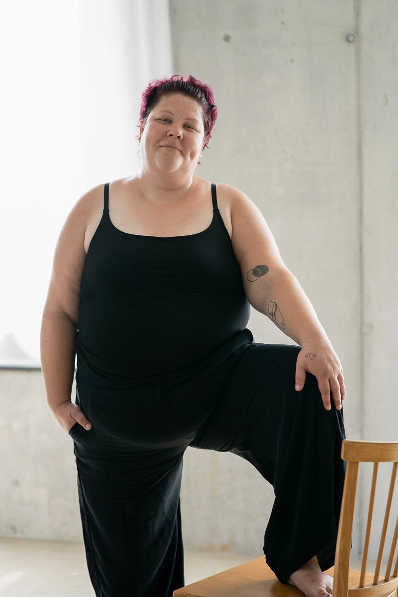 Model steht mit einem Bein auf einem Holzstuhl. Sie trägt das Carla Camitop und die Renée Hose in Schwarz.