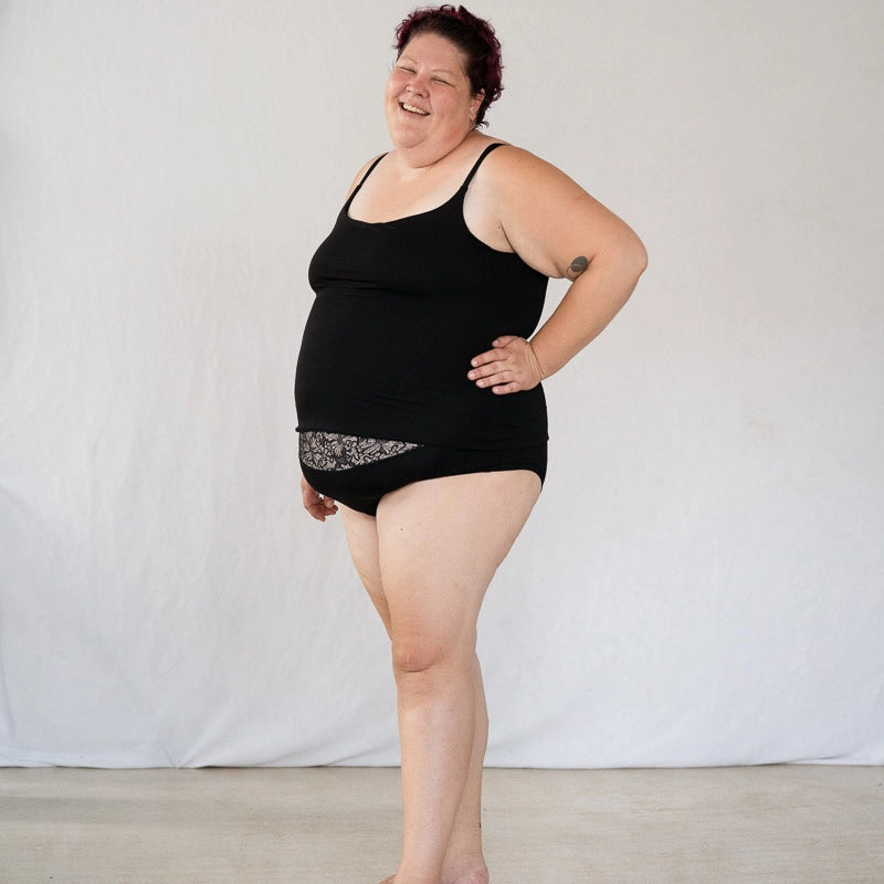 Model steht vor weissem Hintergrund, eine Hand in die Hüfte gestemmt und lächelt in die Kamera. Sie trägt das Carla Camitop und den Nelly Slip in Schwarz.