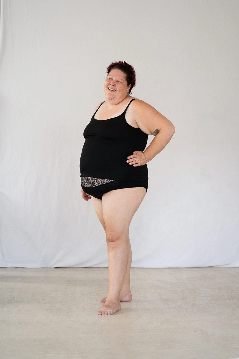 Model steht vor weissem Hintergrund, eine Hand in die Hüfte gestemmt und lächelt in die Kamera. Sie trägt das Carla Camitop und den Nelly Slip in Schwarz.