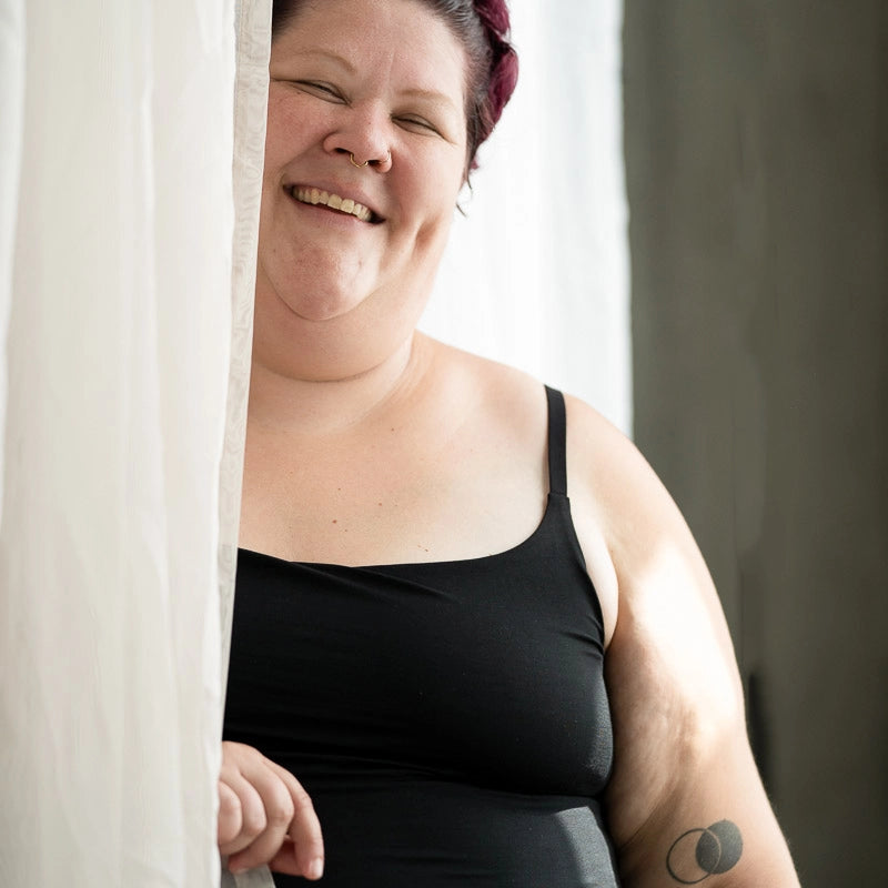 Moodbild, das Model steht beim Fenster am Vorhang und lächelt in die Kamera. Sie tragt das Carla Camitop und den Nelly Slip in Schwarz.