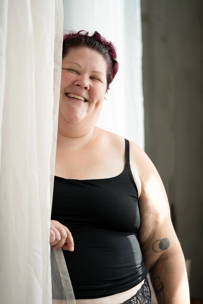 Moodbild, das Model steht beim Fenster am Vorhang und lächelt in die Kamera. Sie tragt das Carla Camitop und den Nelly Slip in Schwarz.