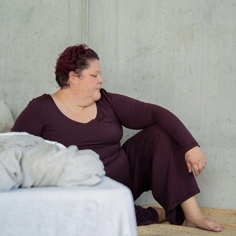 Model sitzt am Boden, seitlich ans Bett gelehnt und blickt zur Seite. Sie trägt das Dolly Longsleeve und die Renée Hose in Aubergine.