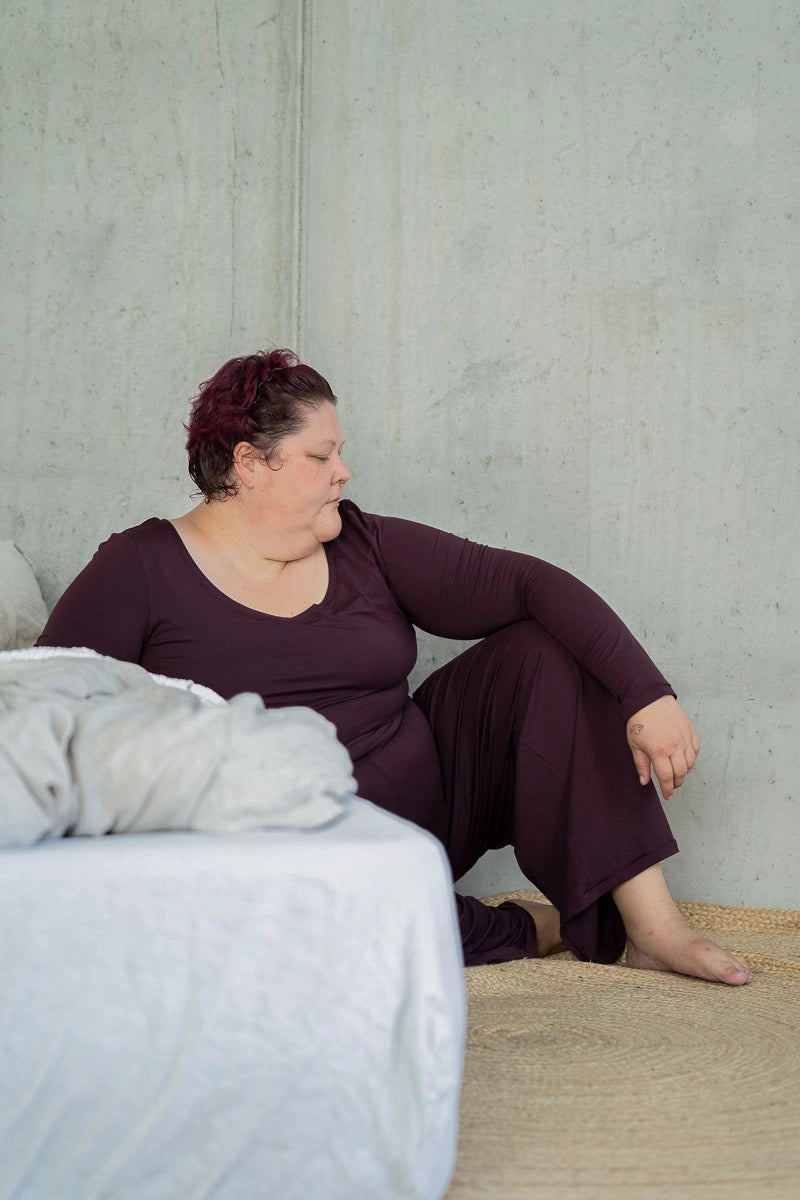 Model sitzt am Boden, seitlich ans Bett gelehnt und blickt zur Seite. Sie trägt das Dolly Longsleeve und die Renée Hose in Aubergine.