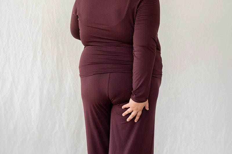 Model steht mit dem Rücken zur Kamera vor weissem Hintergrund. Sie trägt das Dolly Longsleeve und die Renée Hose in Aubergine.