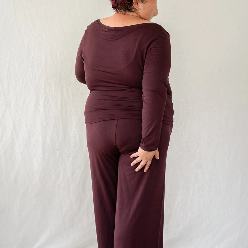 Model steht mit dem Rücken zur Kamera vor weissem Hintergrund. Sie trägt das Dolly Longsleeve und die Renée Hose in Aubergine.
