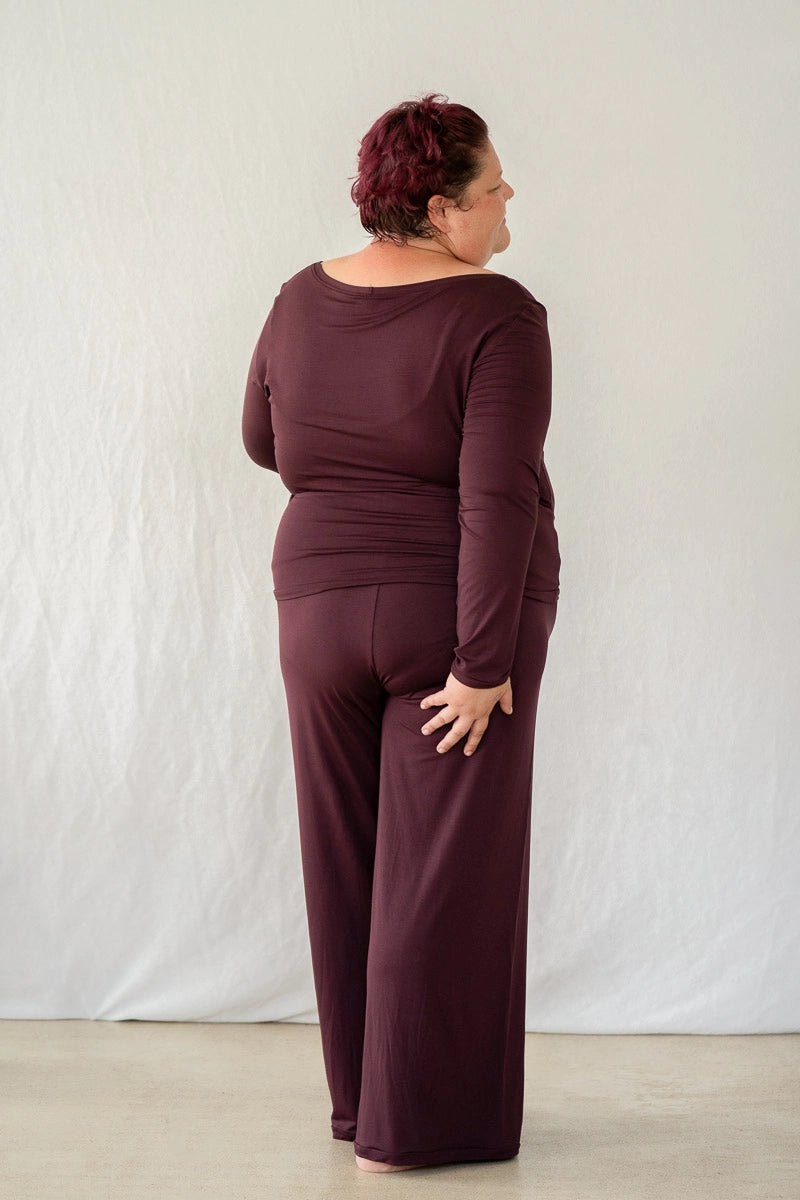 Model steht mit dem Rücken zur Kamera vor weissem Hintergrund. Sie trägt das Dolly Longsleeve und die Renée Hose in Aubergine.