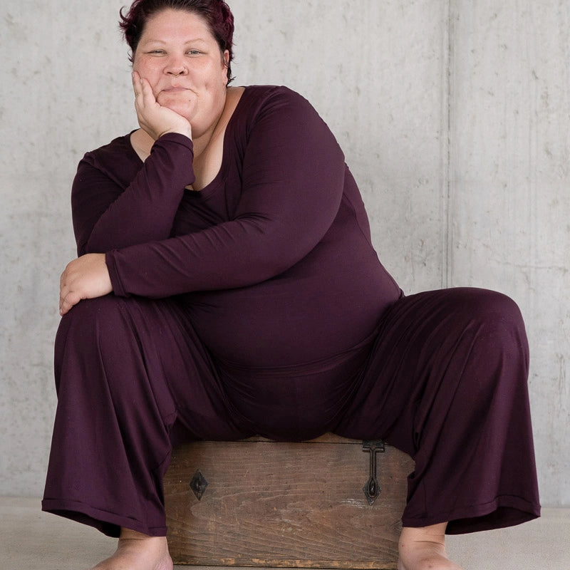 Model sitzt breitbeinig auf einer Holzkiste und grinst in die Kamera. Sie trägt das Dolly Longsleeve und die Renée Hose in Aubergine.