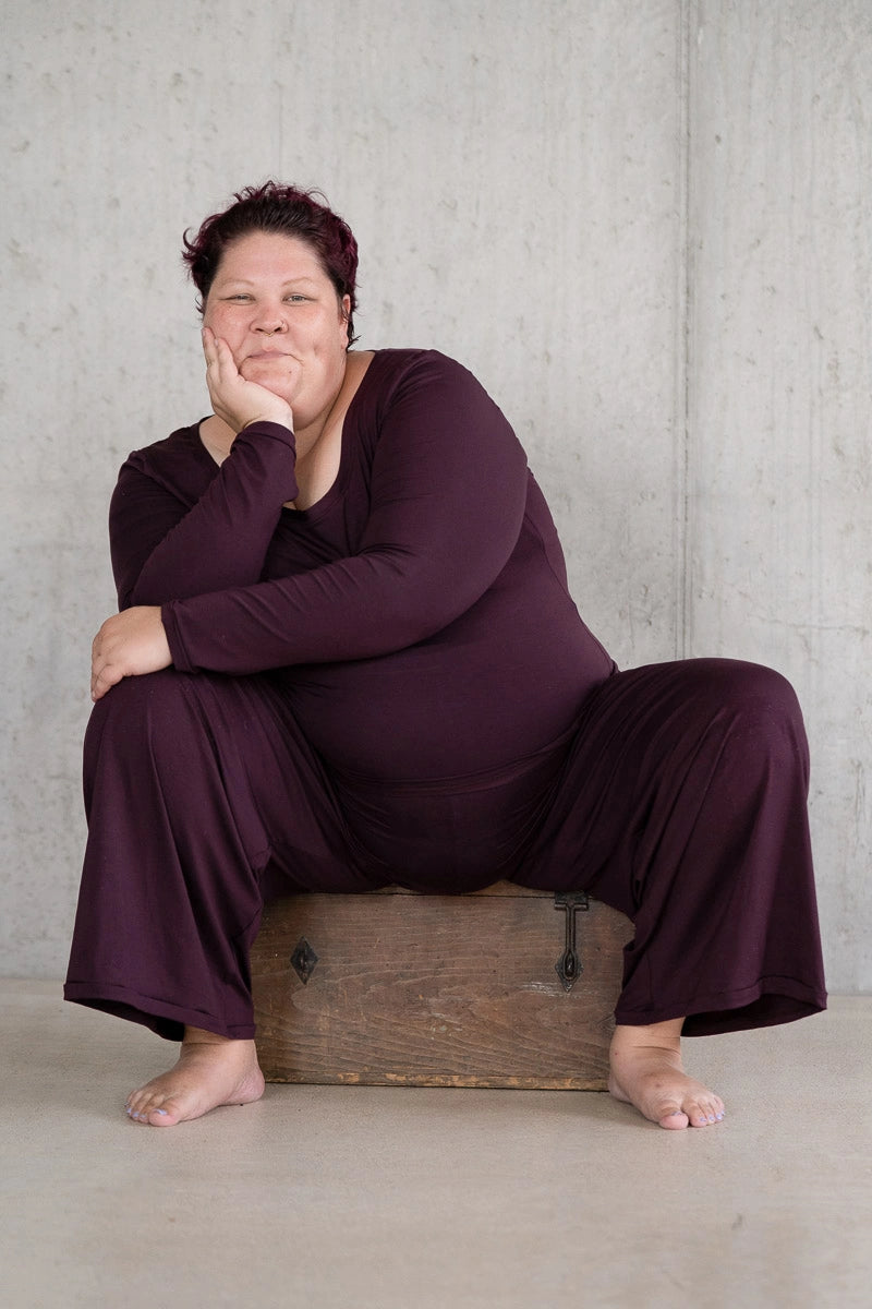 Model sitzt breitbeinig auf einer Holzkiste und grinst in die Kamera. Sie trägt das Dolly Longsleeve und die Renée Hose in Aubergine.