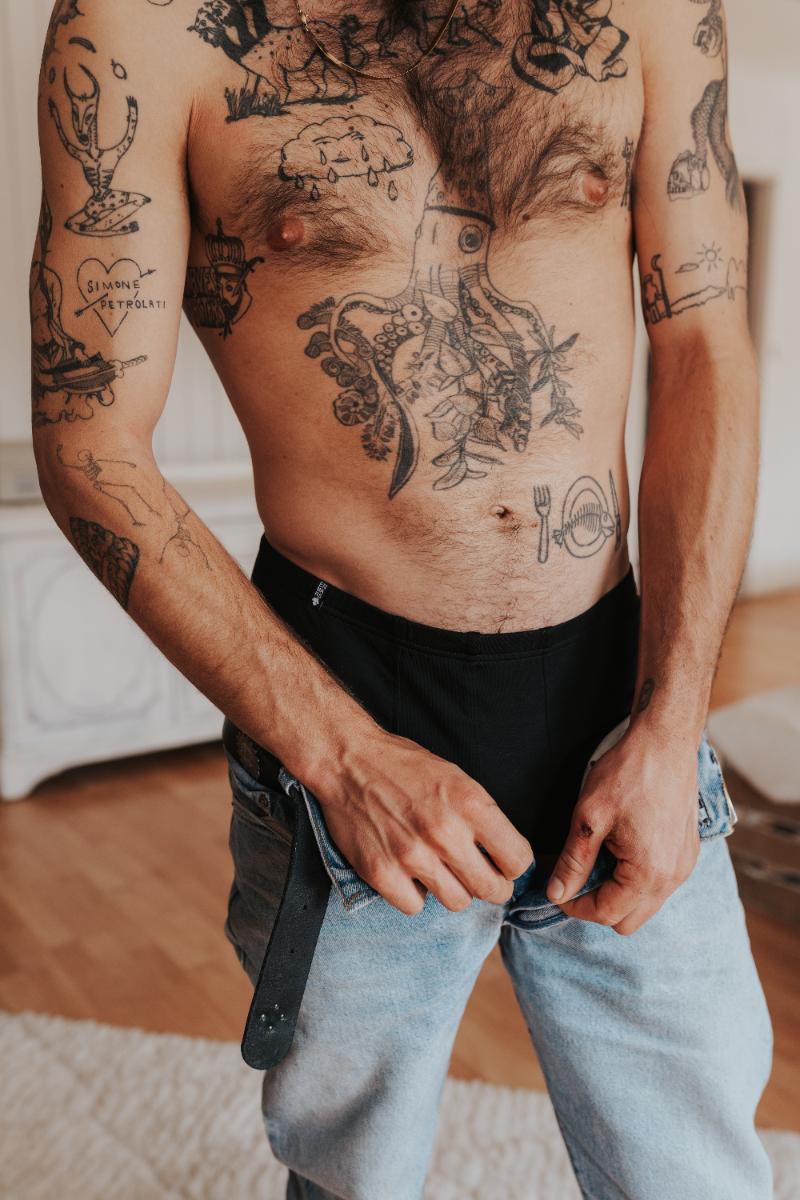 Man with tattoos puts on his pants, under it you can see a black boxer brief.