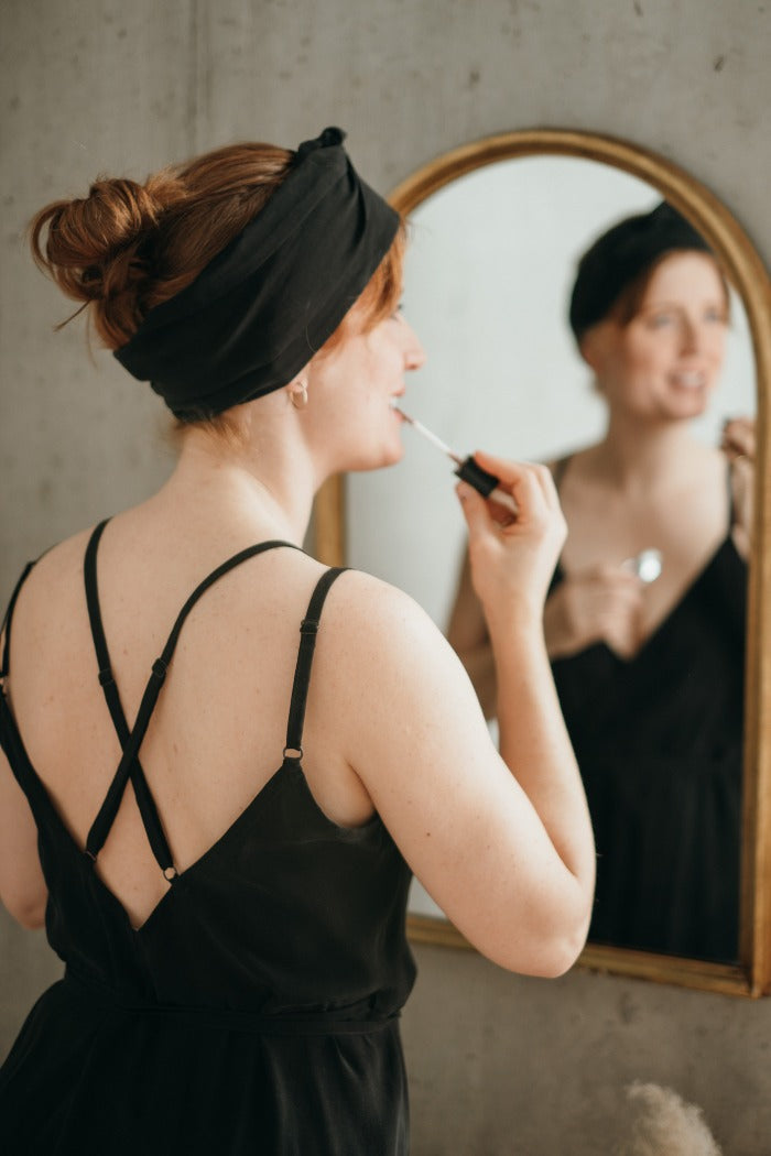 Woman in black dress stands in front of mirror, applies lipstick.