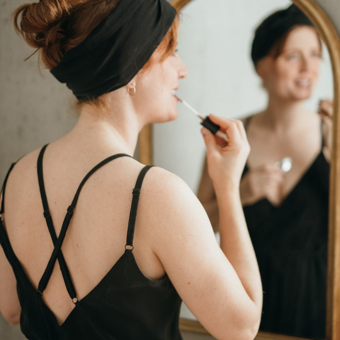 Woman in black dress stands in front of mirror, applies lipstick.