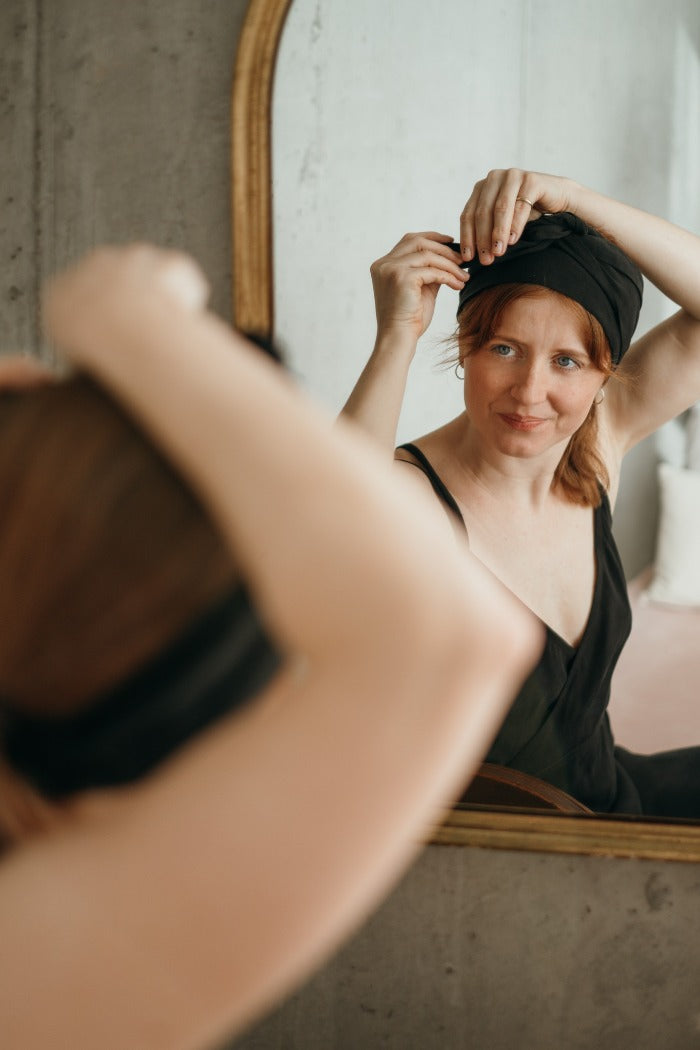 Model in a black slip dress ties a hairband in her hair with concentration.