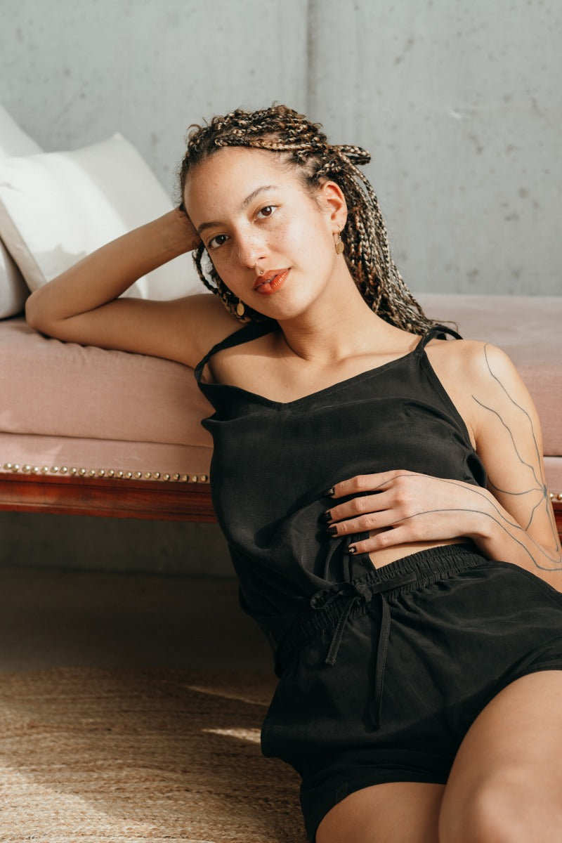 Model leaning against sofa, she wears black top and black shorts.