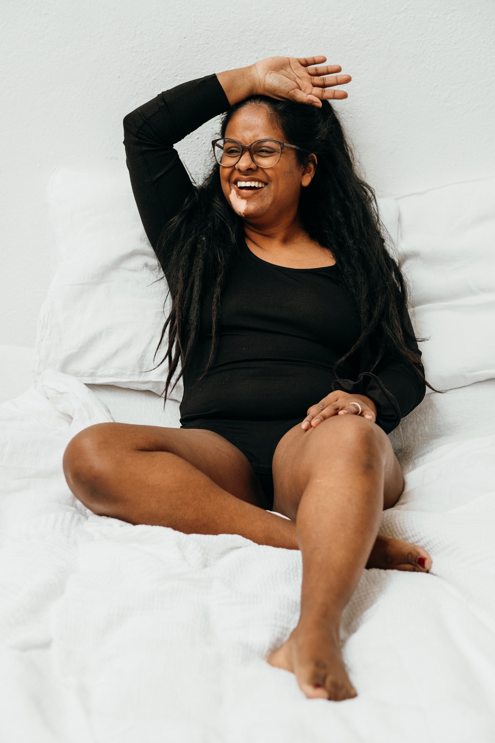 Model trägt schwarzen Langarmbody, sitzt an Wand gelehnt auf dem Bett und lacht.