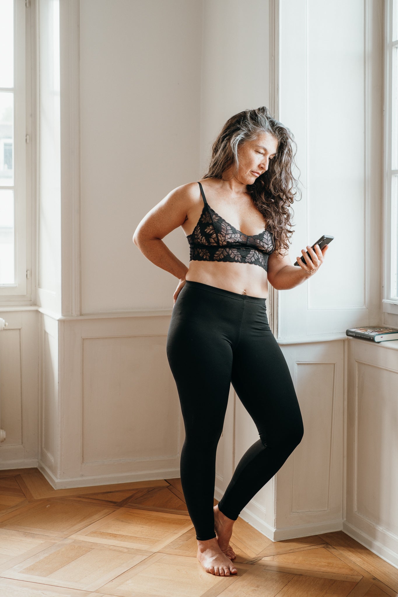 Model leans against wall and looks at her cell phone. She wears leggings and lace bralette.