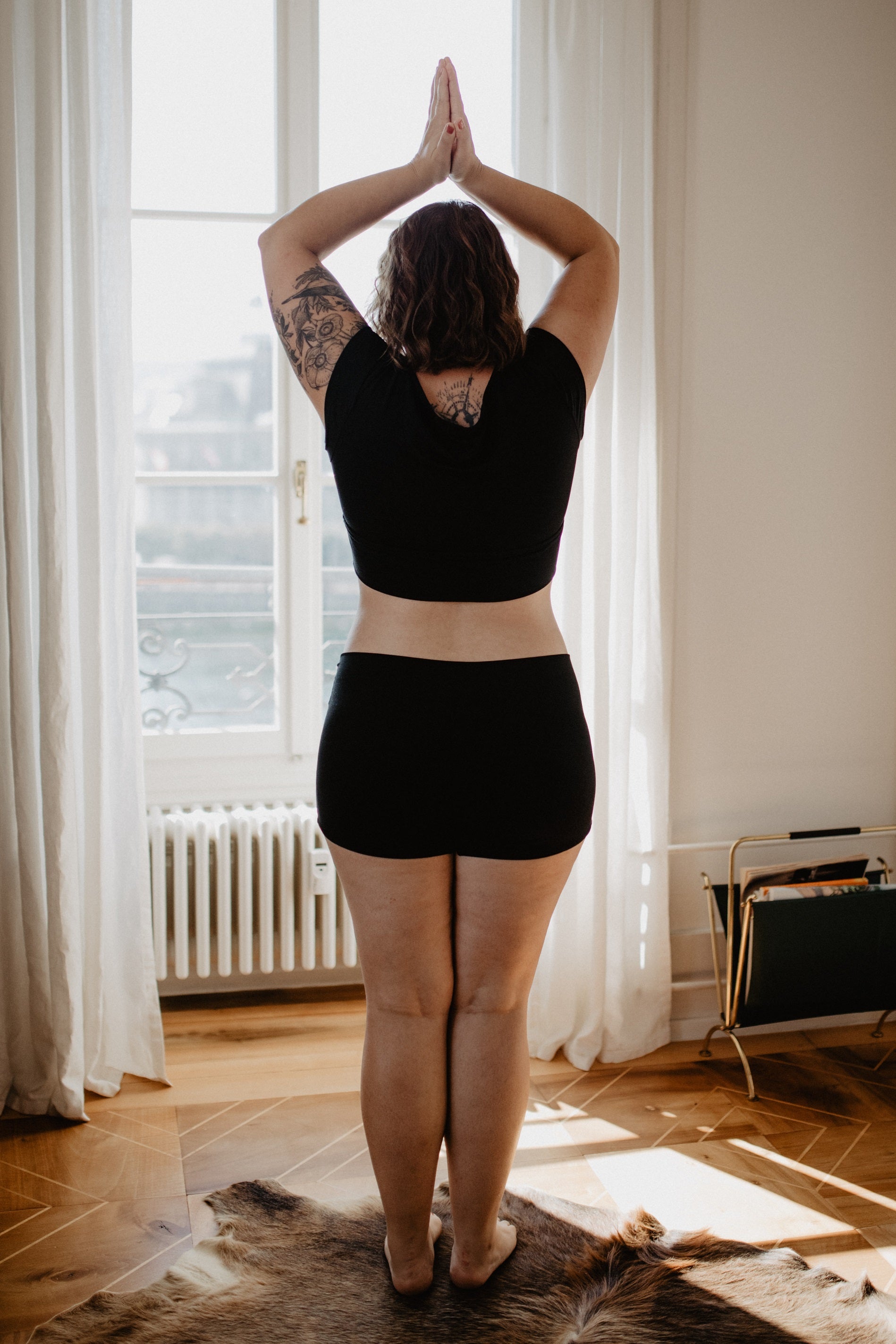 Frau in schwarzem Shirt und Shorts acht Yoga vor dem Fenster.