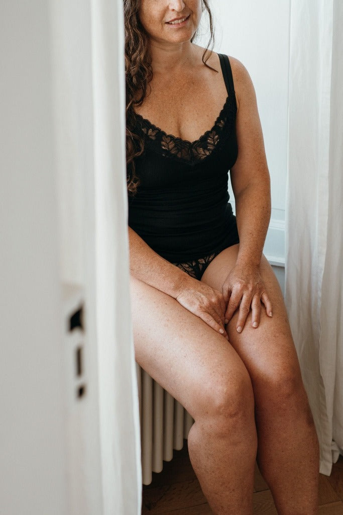 Model sits in front of window, she wears top with lace detail in neckline.